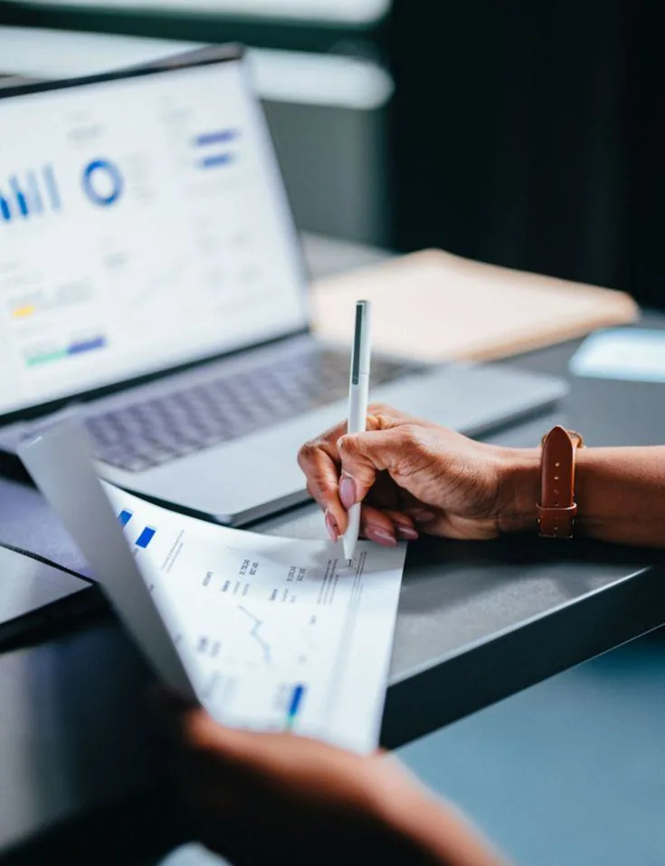 A hand annotating printed financial reports beside a laptop
